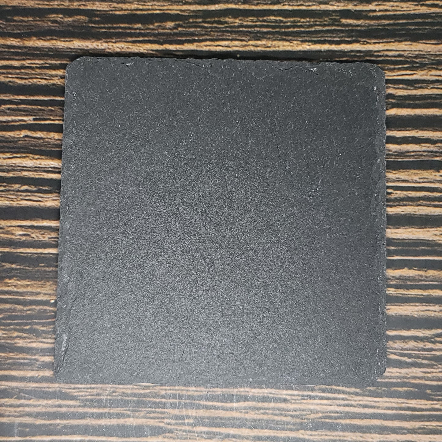 A square black slate tile with rough edges is placed on a wooden surface with a striped pattern. The Custom Slate 4" Coasters - Personalized Laser Engraved Drink Coasters, perfect for custom slate coasters or corporate gifts, is centered in the image and has a matte texture. The wooden surface features a mix of light and dark brown hues. These coasters are brought to you by Insanely Paracord.