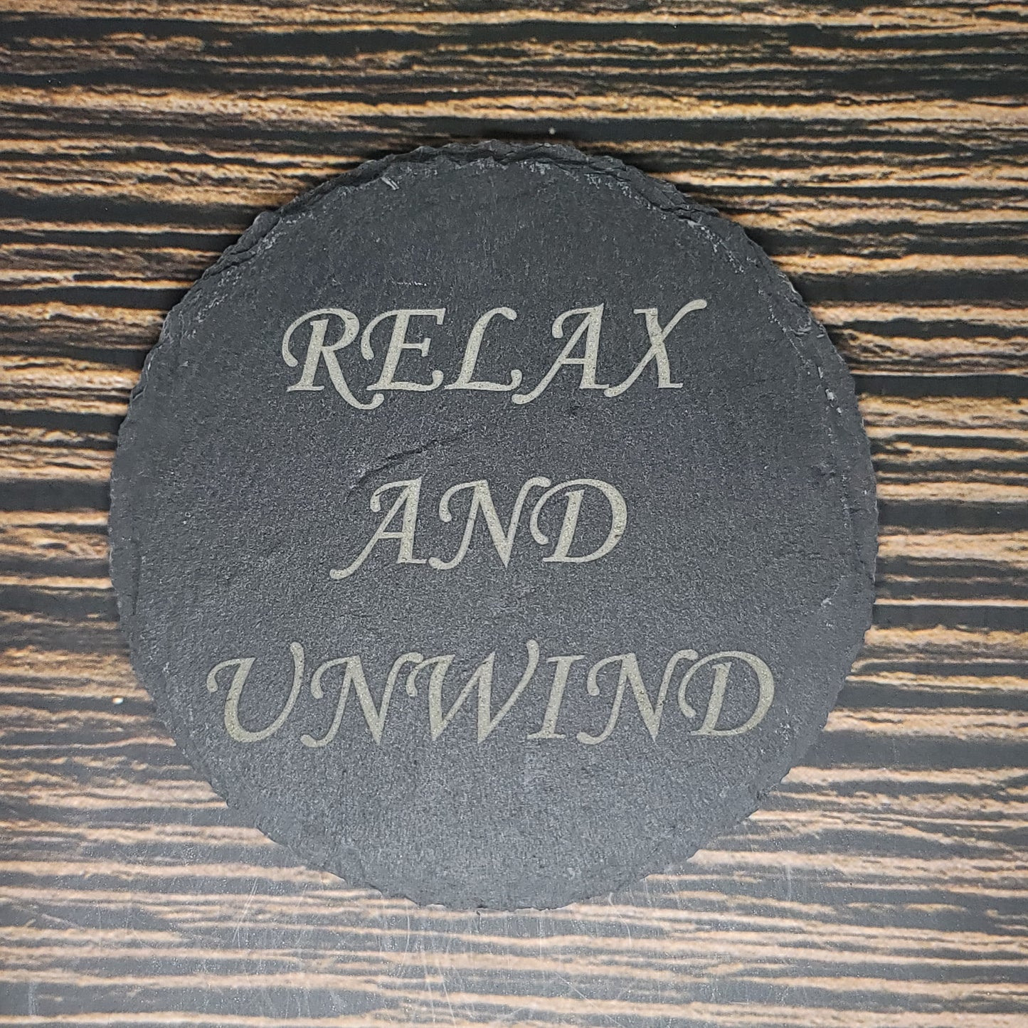 A black, round coaster laser engraved with the words "RELAX AND UNWIND" in white cursive text sits against a dark wooden background with horizontal grain lines. Perfect as a unique corporate gift, these Custom Slate 4" Coasters - Personalized Laser Engraved Drink Coasters by Insanely Paracord add a touch of elegance to any setting.