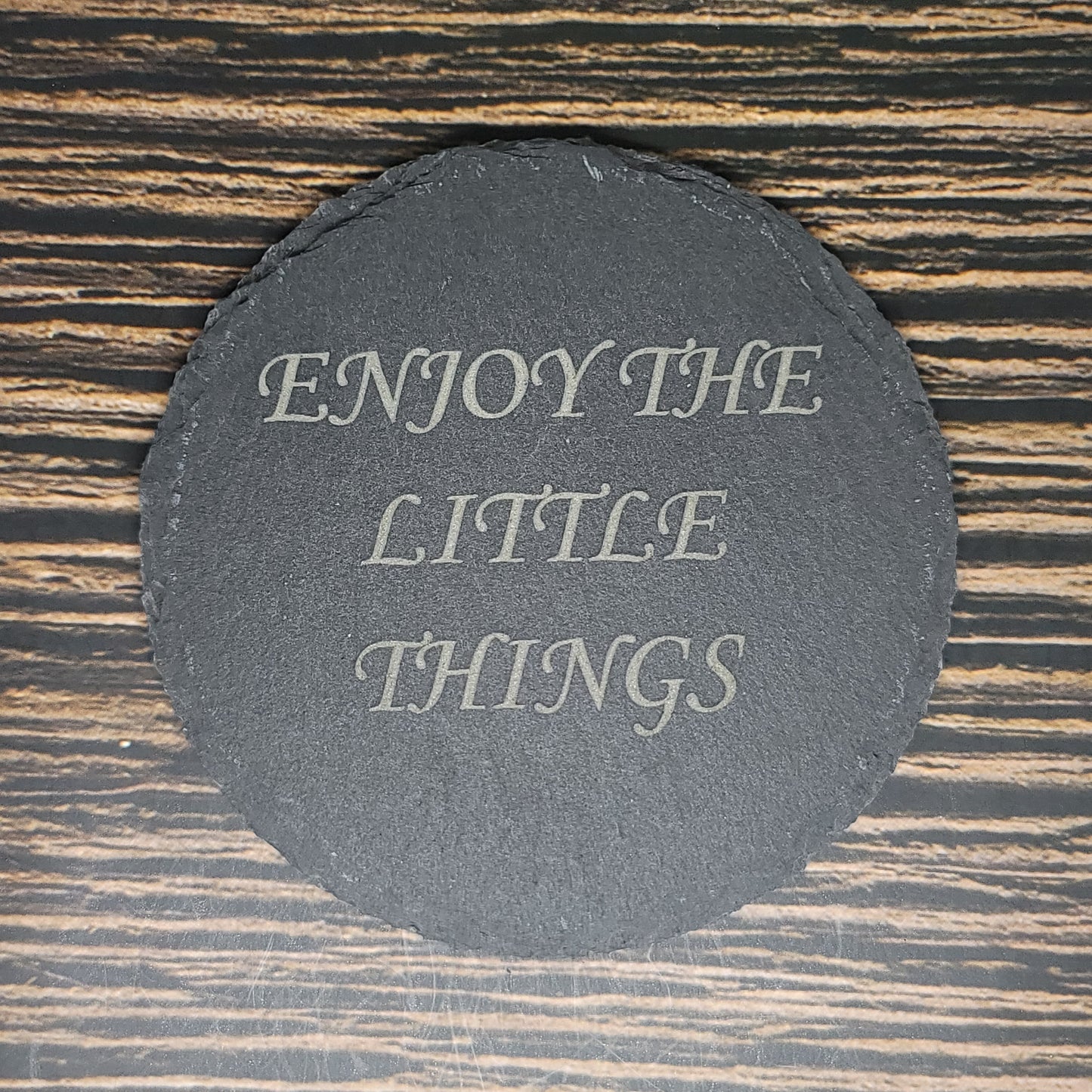 A round, black Custom Slate 4" Coaster from Insanely Paracord with the text "ENJOY THE LITTLE THINGS" written in elegant white letters is placed on a wooden surface with dark and light brown stripes.