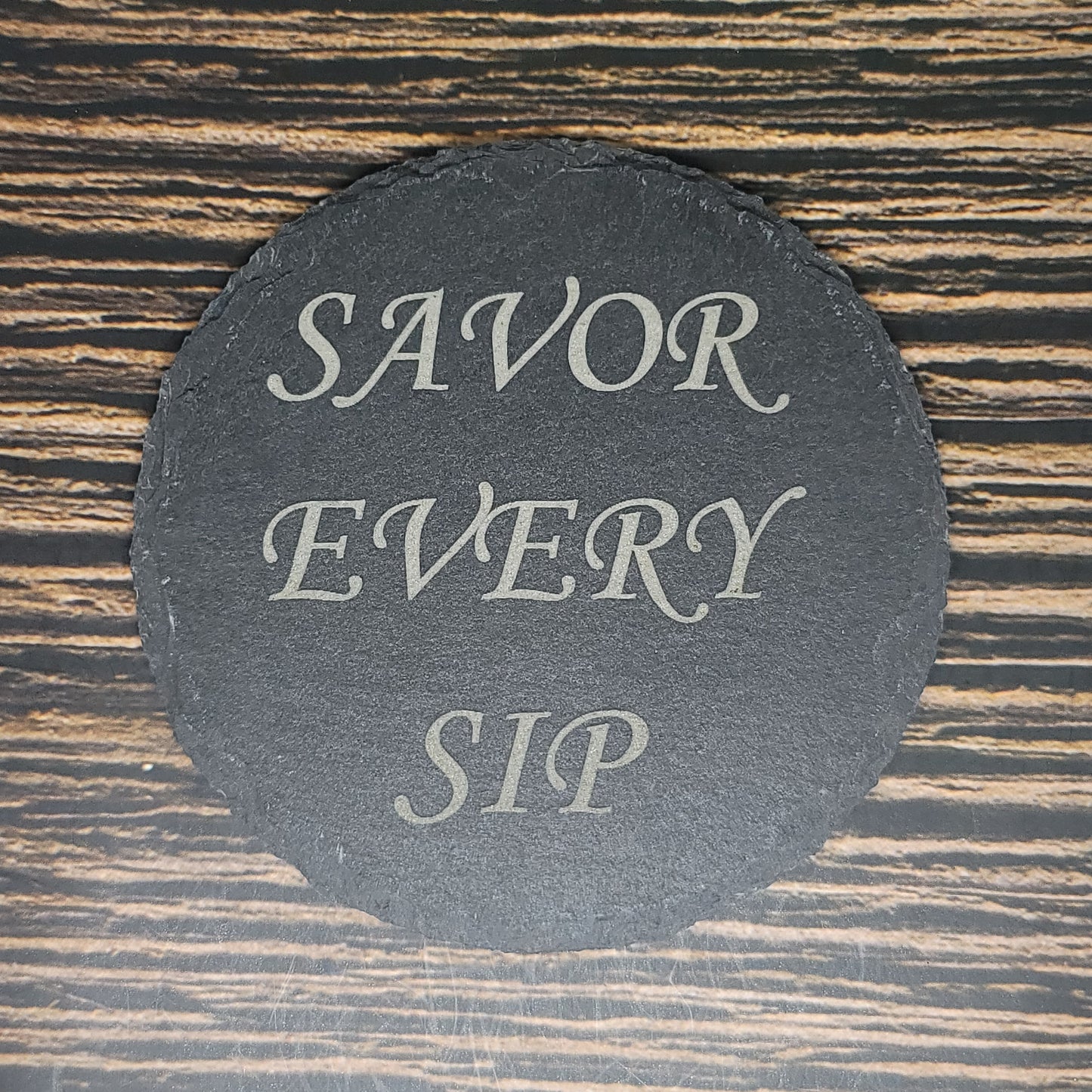 A circular, custom slate coaster with the text "SAVOR EVERY SIP" written in elegant, light-colored script is placed on a wooden surface with horizontal grain patterns. Ideal as a corporate gift, this stylish addition enhances any setting. These Custom Slate 4" Coasters - Personalized Laser Engraved Drink Coasters by Insanely Paracord perfectly blend functionality and aesthetics.