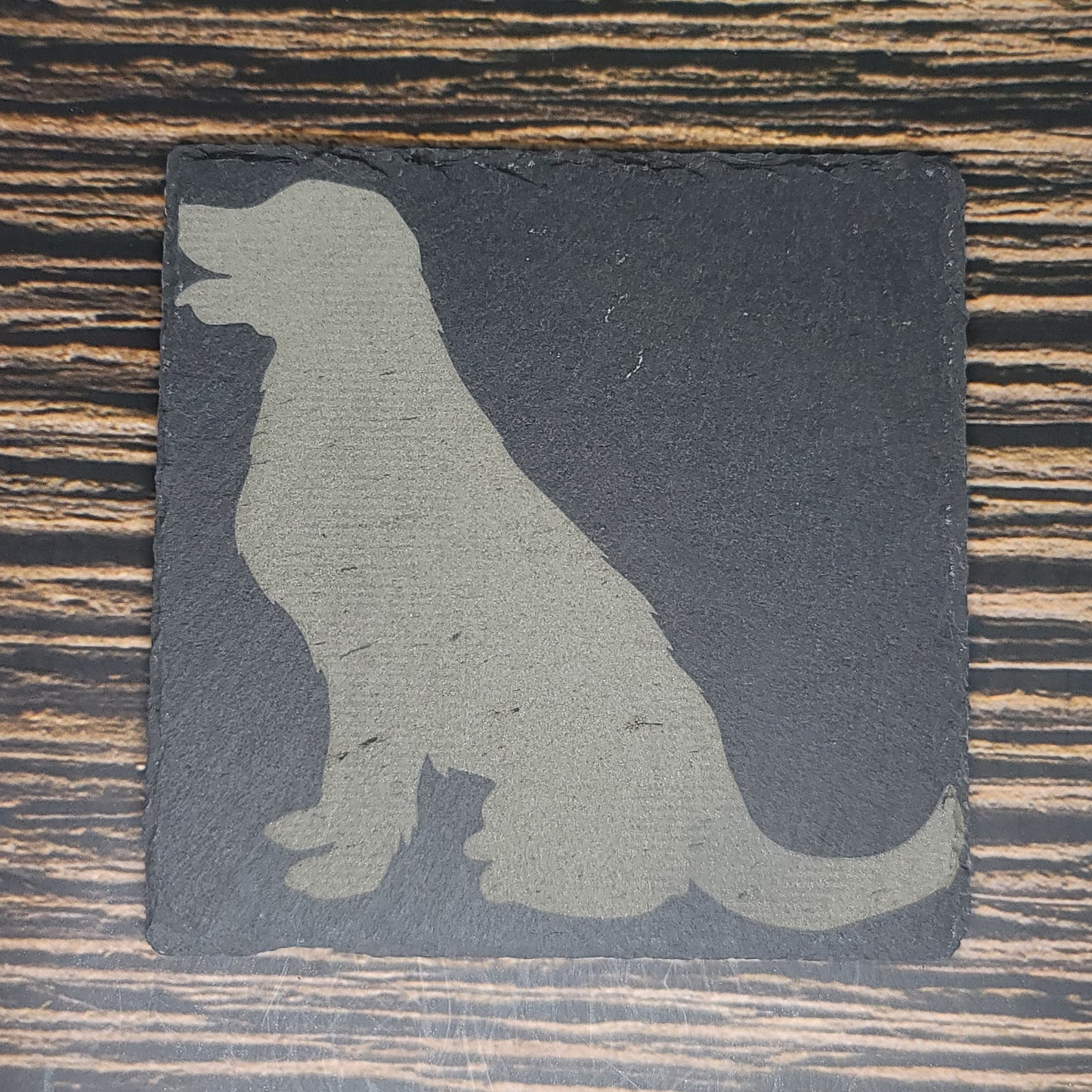 A Custom Slate 4" Coaster - Personalized Laser Engraved Drink Coaster by Insanely Paracord with a silhouette of a sitting dog laser engraved on it. The dog is facing left with its mouth slightly open. The textured surface of the coaster complements the wooden table's visible grain patterns, making it an ideal corporate gift.