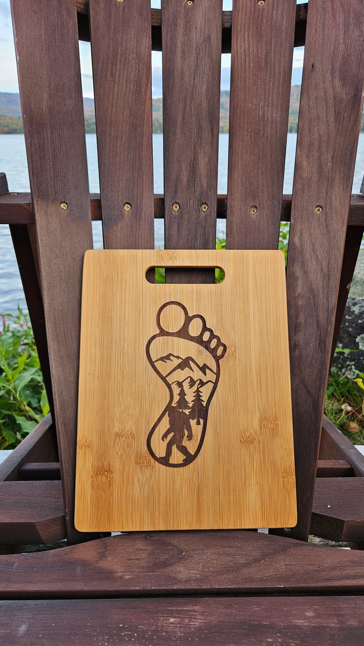A laser-engraved **Lake Pleasant Bamboo (12x8) or Acacia (12x9) Cutting Board** by **Insanely Paracord** with a footprint design featuring a mountain and forest landscape is placed on a wooden chair. In the background, there is a lake with trees and hills visible on the other side. This custom cutting board makes an ideal corporate gift.