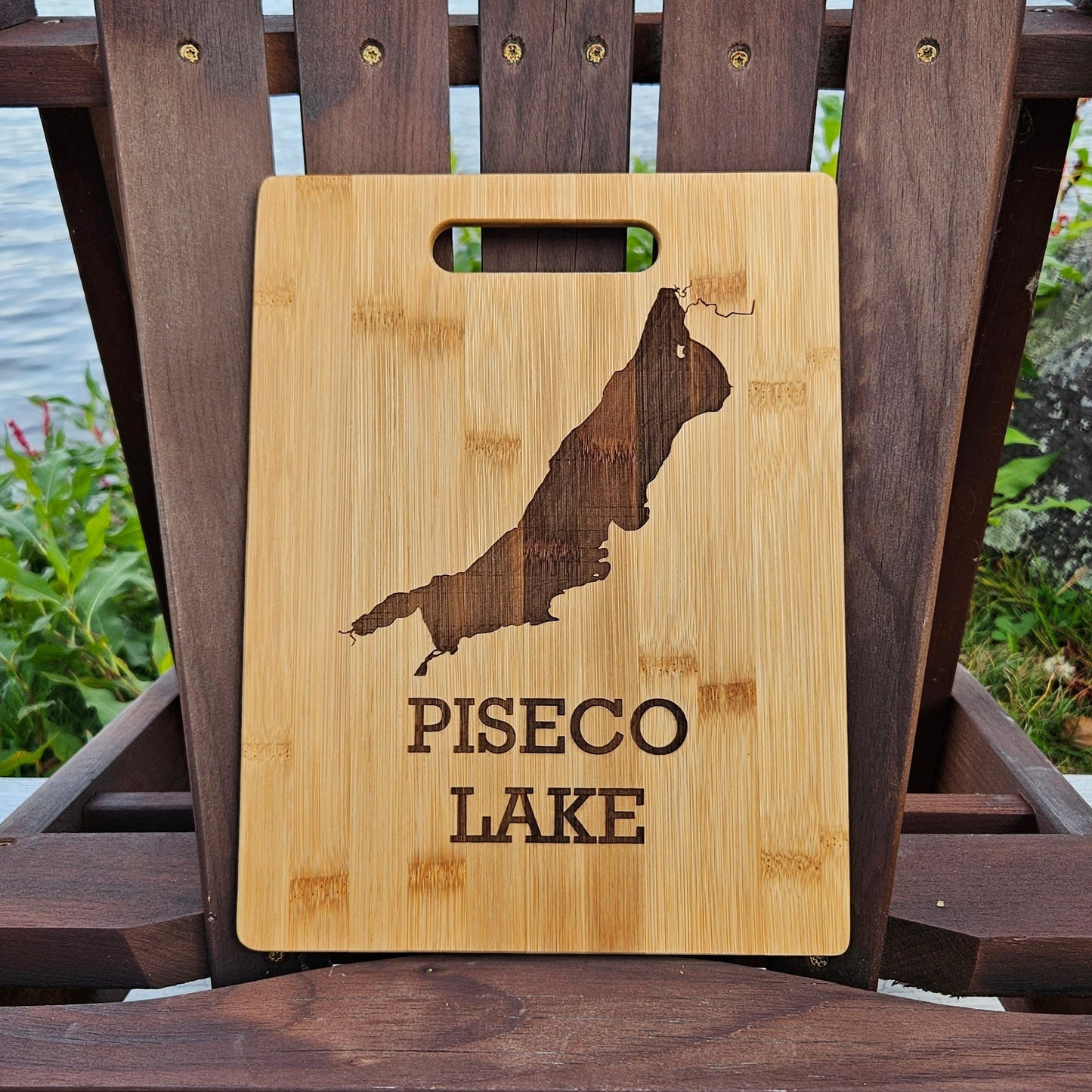 A beautifully laser engraved wooden cutting board, featuring the outline of Piseco Lake and the text "PISECO LAKE," is showcased on an Adirondack chair. The chair sits outdoors, with lush greenery and a serene lake in the background. This custom Insanely Paracord Piseco Lake Bamboo (12x8) or Acacia (12x9) Cutting Board makes for a perfect corporate gift.