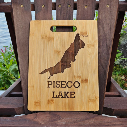 A beautifully laser engraved wooden cutting board, featuring the outline of Piseco Lake and the text "PISECO LAKE," is showcased on an Adirondack chair. The chair sits outdoors, with lush greenery and a serene lake in the background. This custom Insanely Paracord Piseco Lake Bamboo (12x8) or Acacia (12x9) Cutting Board makes for a perfect corporate gift.
