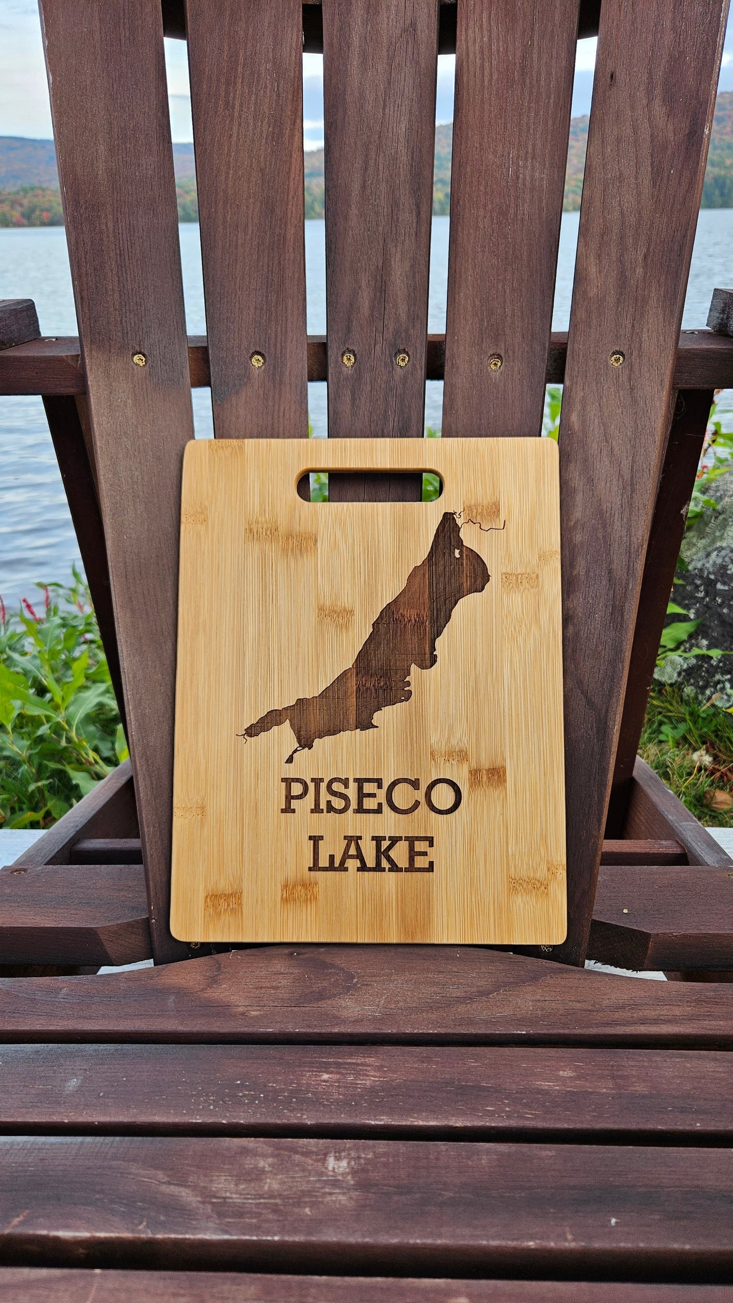 A Lake Pleasant Bamboo (12x8) or Acacia (12x9) Cutting Board by Insanely Paracord with a laser-engraved image of a lake and the text "PISECO LAKE" sits upright on a wooden chair. The chair is positioned outdoors with a calm lake and lush foliage in the background, making it an ideal corporate gift.