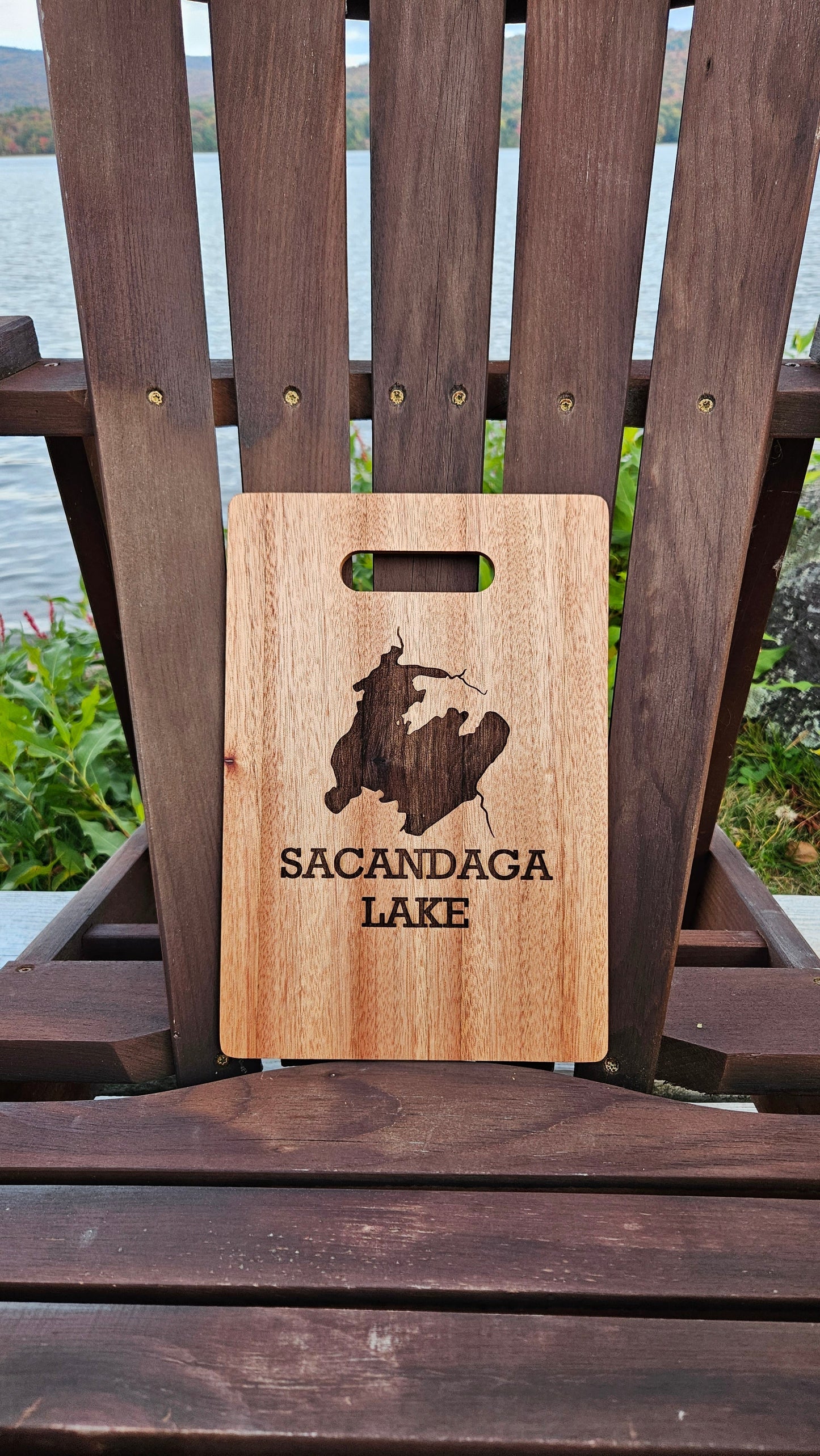 An Insanely Paracord Lake Pleasant Bamboo (12x8) or Acacia (12x9) Cutting Board with a handle, featuring the engraving of a bear and the text "Sacandaga Lake," is placed on a wooden chair. In the background, there is a lake and some greenery, making it a perfect corporate gift.