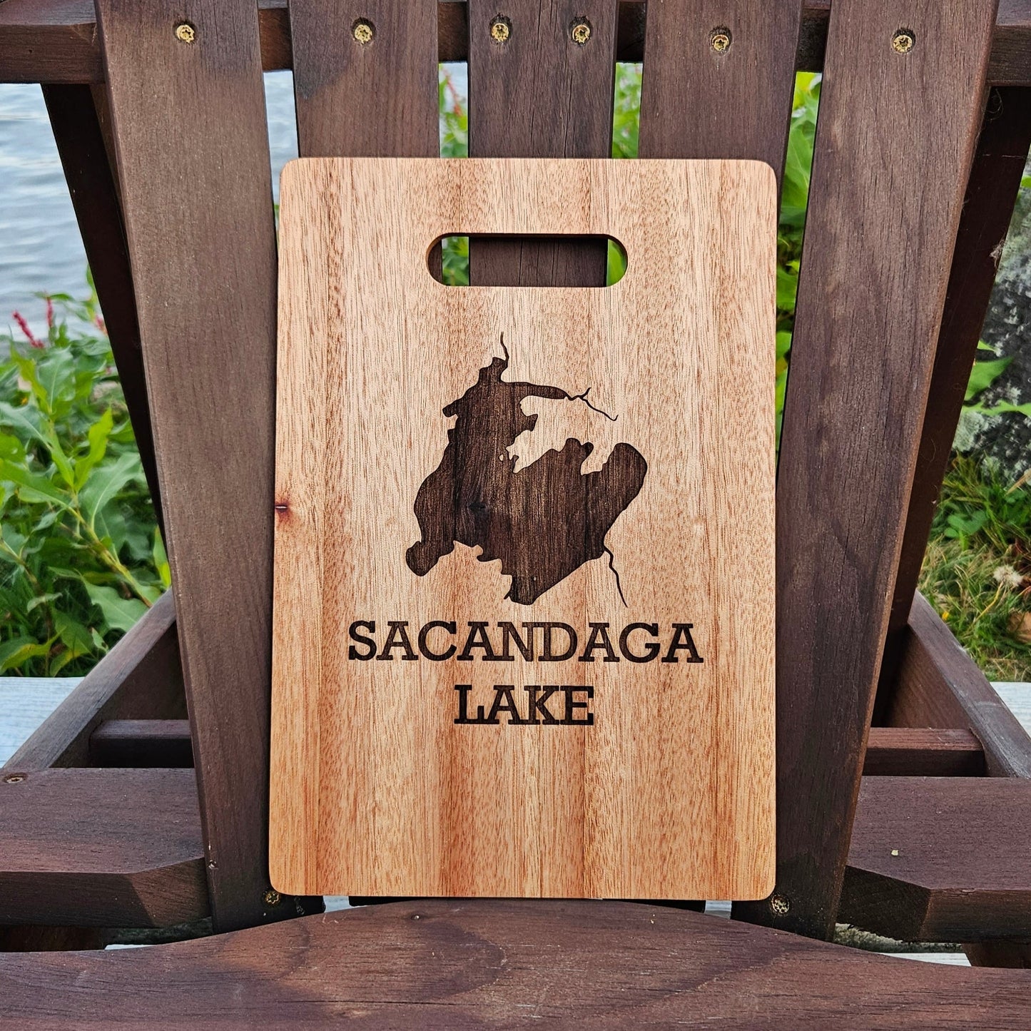 An Insanely Paracord Sacandaga Lake Bamboo (12x8) or Acacia (12x9) Cutting Board is placed on a dark brown wooden chair by a lake. The custom cutting board features a laser engraved image of a lake map with the words "SACANDAGA LAKE" underneath. In the background, there are green plants and water, making it the perfect personalized gift.