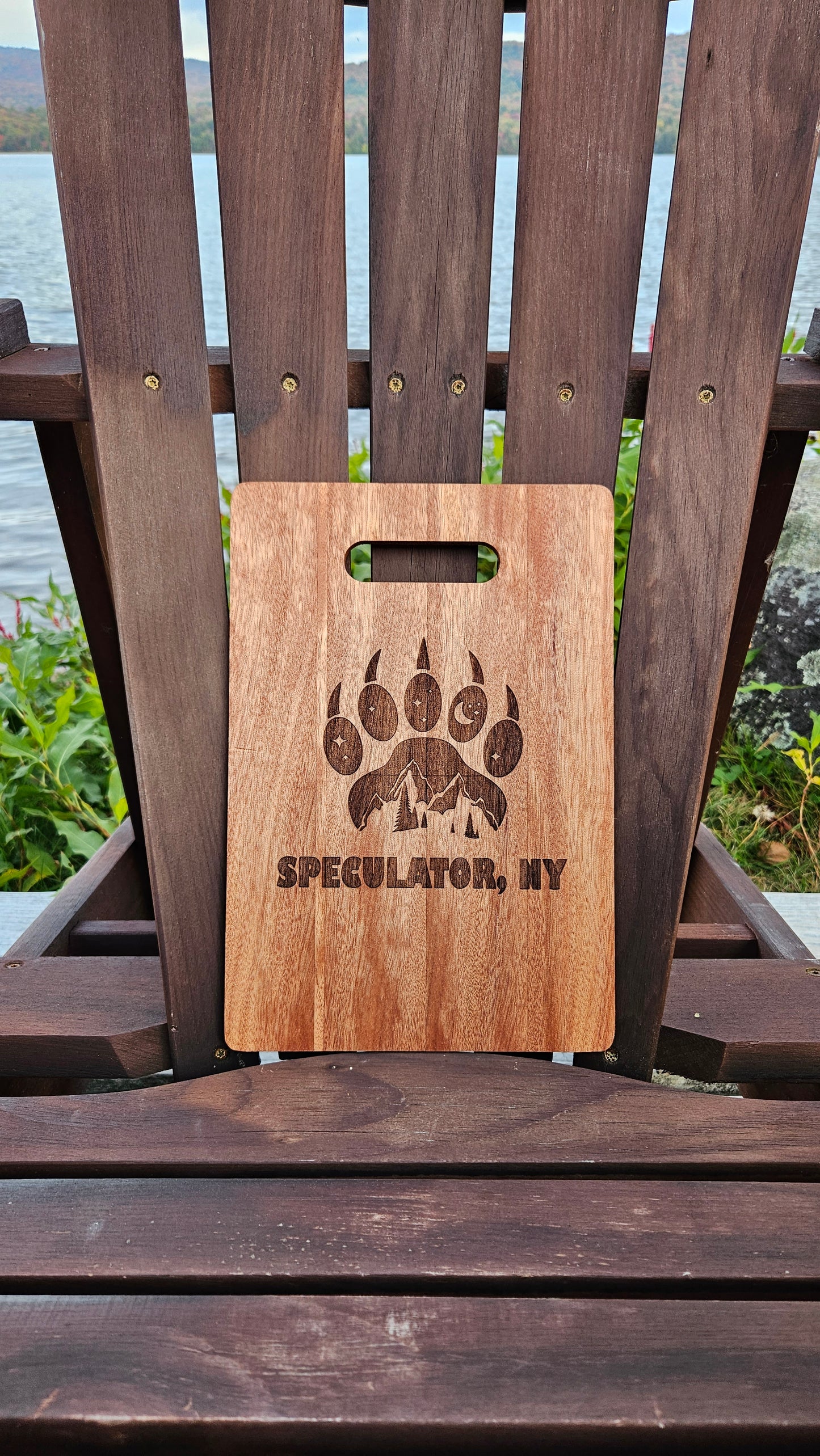 A Custom Bamboo (12x8) or Acacia (12x9) Cutting Board by Insanely Paracord with the text "SPECULATOR, NY" and an engraved design featuring mountains and a bear paw is placed on a wooden Adirondack chair. The laser-engraved piece sits near a lake with hills and foliage in the background, making it an ideal corporate gift.