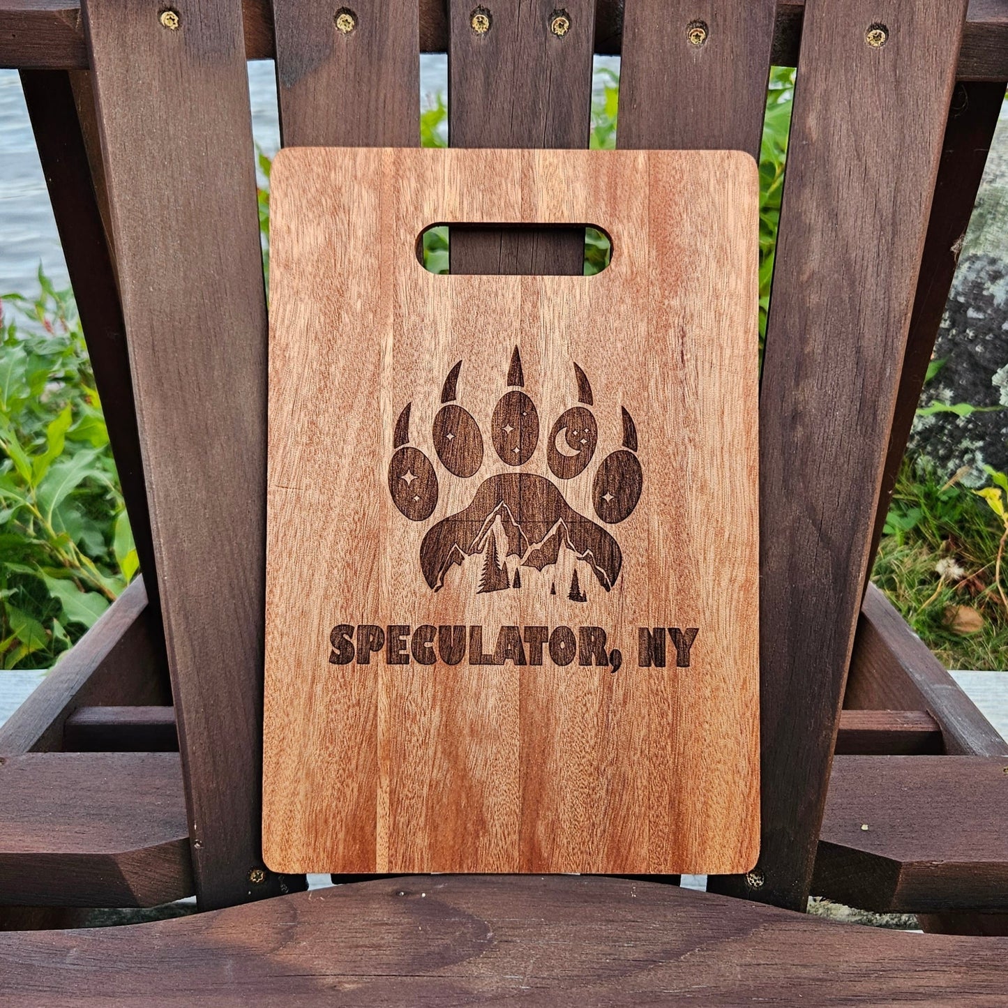 An Insanely Paracord Speculator, NY Bamboo (12x8) or Acacia (12x9) Cutting Board is placed on an Adirondack chair outdoors. The laser engraved cutting board features a design of a bear paw with mountains inside it and the text "SPECULATOR, NY" beneath the design. There is greenery and water in the background, making it perfect for personalized gifts.