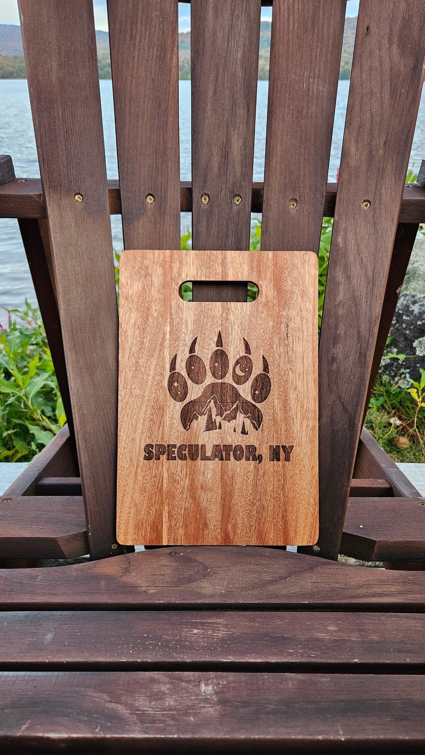 A Lake Pleasant Bamboo (12x8) or Acacia (12x9) Cutting Board by Insanely Paracord, with a handle, laser engraved with a bear paw print and pine trees. The text below reads "SPECULATOR, NY." This custom cutting board, ideal as a corporate gift, rests on a wooden chair by a lake, surrounded by lush greenery under a cloudy sky.