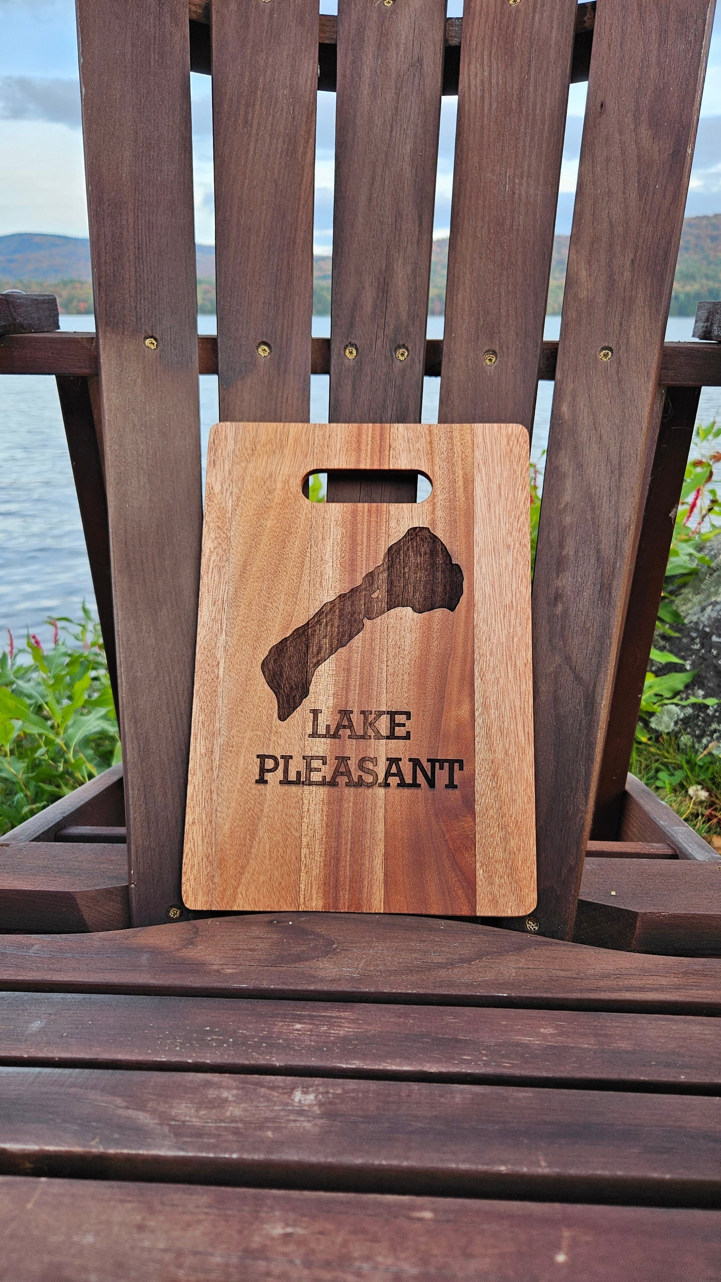 A Sacandaga Lake Bamboo (12x8) or Acacia (12x9) Cutting Board by Insanely Paracord, laser engraved with "Lake Pleasant" and an outline of the lake, is placed on an Adirondack chair by the waterfront. With green foliage and mountains in the background under a cloudy sky, it's a perfect scene for personalized gifts or custom cutting boards.