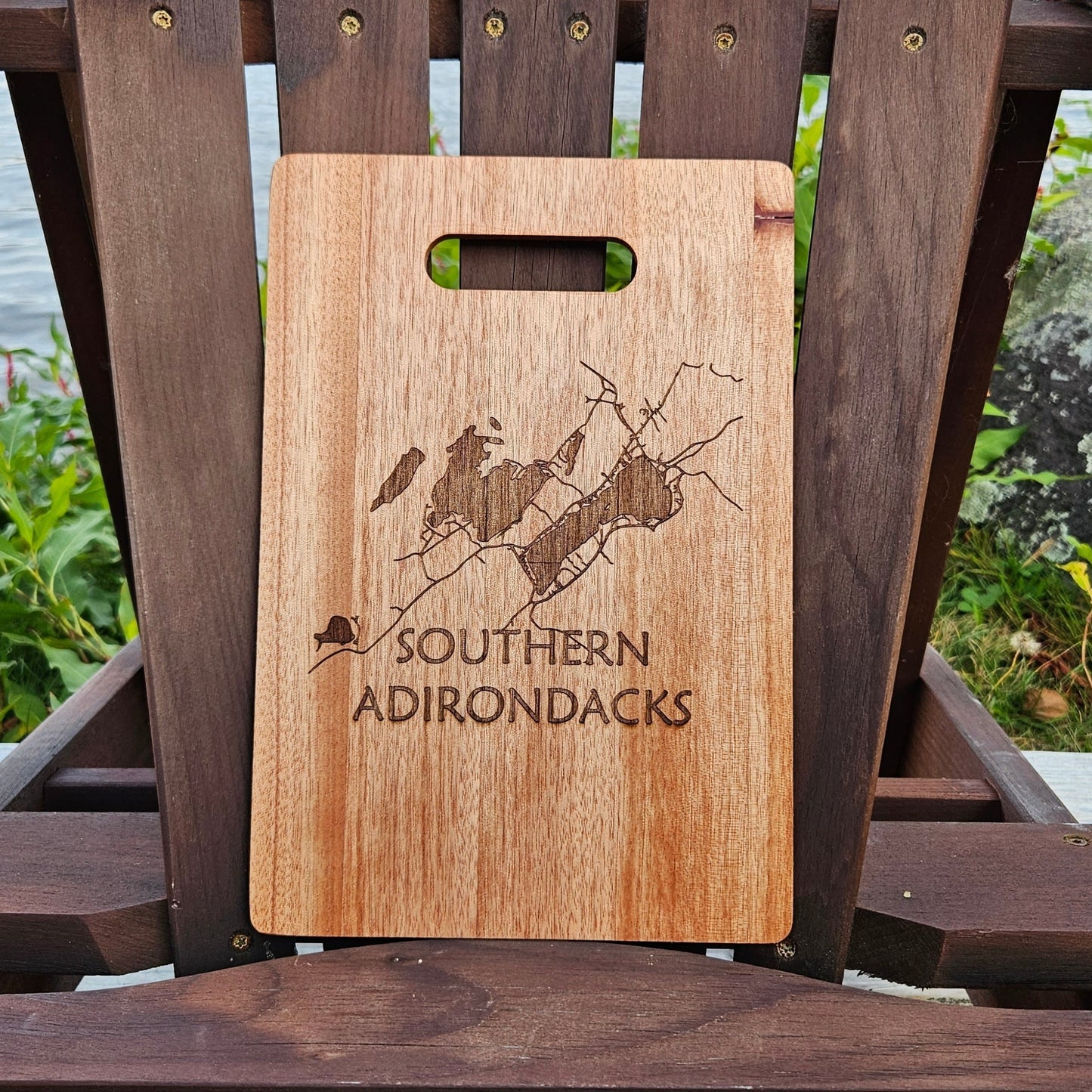 A Southern Adirondacks Bamboo (12x8) or Acacia (12x9) Cutting Board by Insanely Paracord, laser engraved with a map and the text "Southern Adirondacks," sits on an Adirondack chair. The background includes lush greenery and a serene body of water, suggesting an outdoor, lakeside location—perfect for custom cutting boards or unique laser engraved gifts.