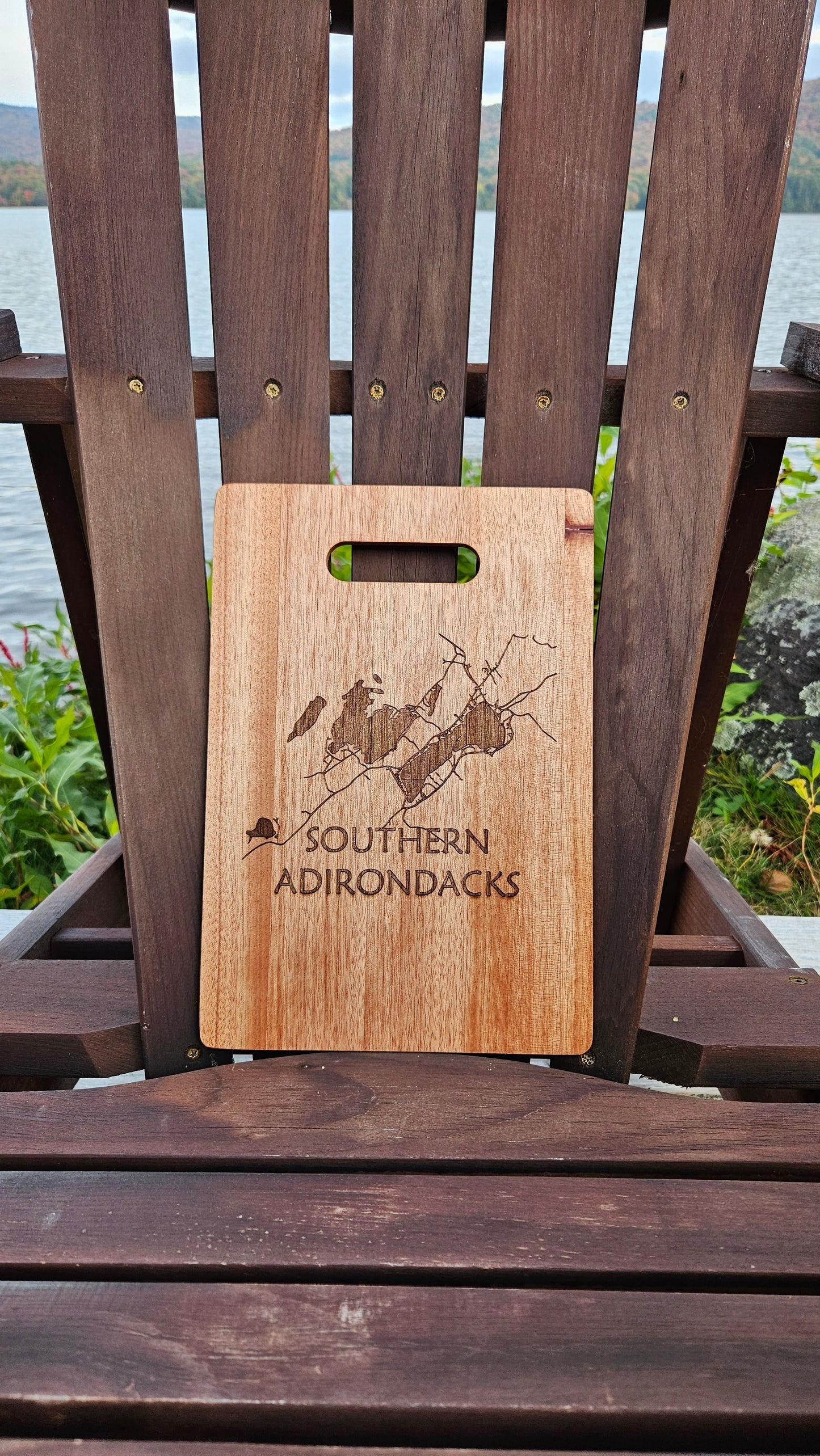 A Sacandaga Lake Bamboo (12x8) or Acacia (12x9) Cutting Board by Insanely Paracord with a laser-engraved map and the words "Southern Adirondacks" rests against the back of a wooden chair. The chair is positioned outdoors by a lake, framed by greenery and mountains in the background, making it an ideal setting for personalized gifts.