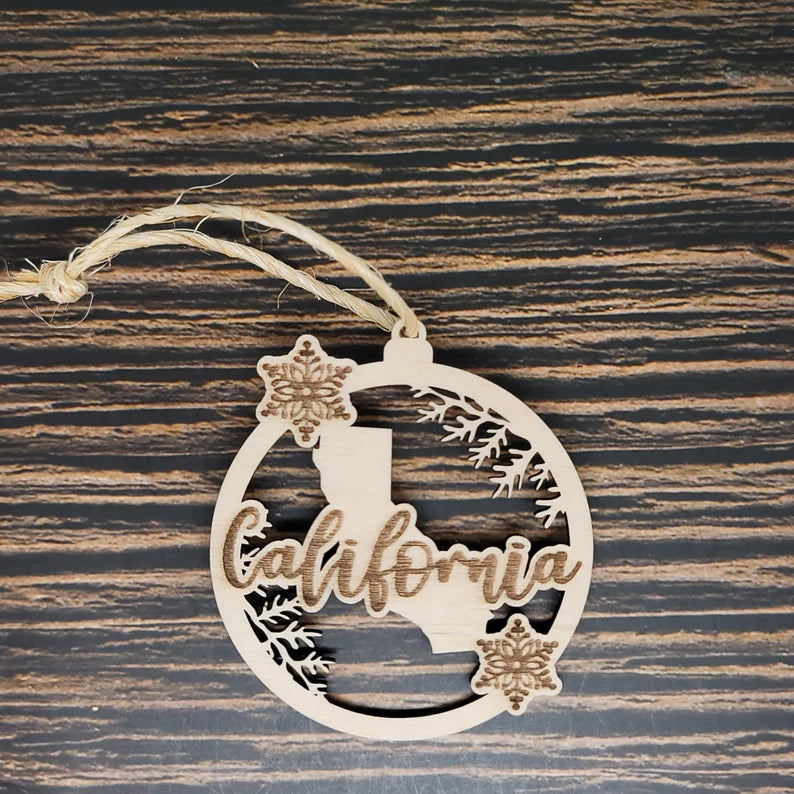 A California State Christmas Ornament - Laser Engraved Birch Wood by Insanely Paracord featuring the silhouette of California with "California" elegantly cut out in a script font. This enchanting piece is decorated with two snowflakes and delicate branches, laser engraved for precision, and hangs from rustic twine on a dark wooden background.