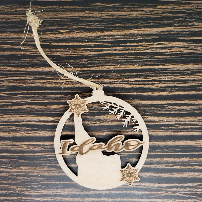 An Insanely Paracord Idaho State Laser Engraved Christmas Ornament - 3" Tall Birch Wood Tree Decoration featuring a laser-engraved scene of Idaho. The ornament has a circular shape with a cutout depicting the state's outline, along with snowflakes and decorative branches. A twine loop is attached at the top for hanging.