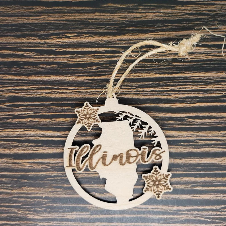 A rustic holiday decor piece, the Illinois Wooden State Christmas Ornament - Laser Engraved Birch Wood - 3" Tall, 2.75" Wide by Insanely Paracord features the outline of Illinois with "Illinois" scripted across it. This laser-engraved birch wood ornament is circular with snowflake designs and a twine loop for hanging, all set on a dark wood surface.