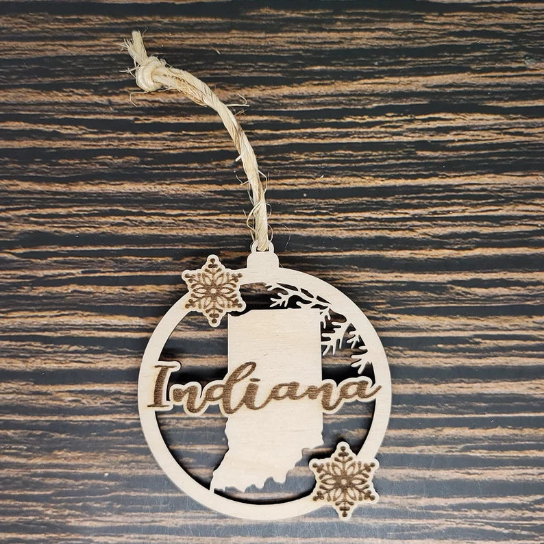 An Insanely Paracord Indiana State Laser Engraved Christmas Ornament - 3" x 2.75" Birch Wood features the word "Indiana" with an outline of the state in the background. Two snowflakes and a small branch decorate the circular Indiana State Laser Engraved Christmas Ornament - 3" x 2.75" Birch Wood. It's hanging by a braided rope against a wood-textured surface.