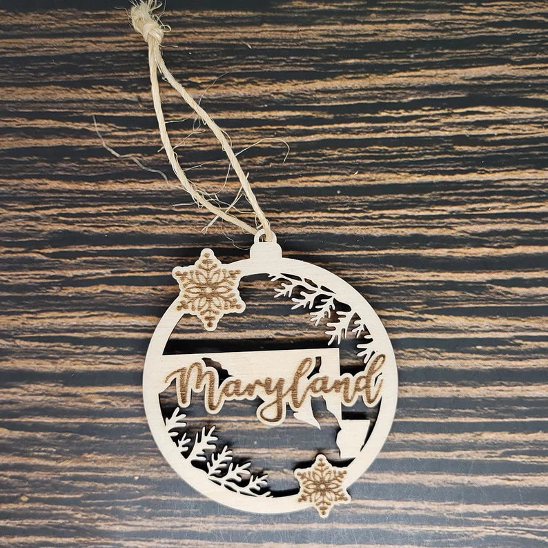 An Insanely Paracord Maryland State Christmas Ornament - Laser Engraved Birch Wood - 3" Tall - Natural Twine, features the state's outline adorned with snowflakes and pine branches. The word "Maryland" is carved across the middle. The ornament hangs from a twine loop and rests on a dark-textured wooden surface.