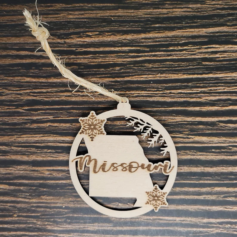 A wooden Christmas ornament featuring the outline of Missouri with "Missouri" stylishly engraved across it. This Missouri State Laser Engraved Christmas Ornament - 3" Tall, 2.75" Wide, Birch Wood by Insanely Paracord is decorated with two snowflakes and a pine branch, hanging by a rustic twine loop against a wooden background, perfect for holiday decor.
