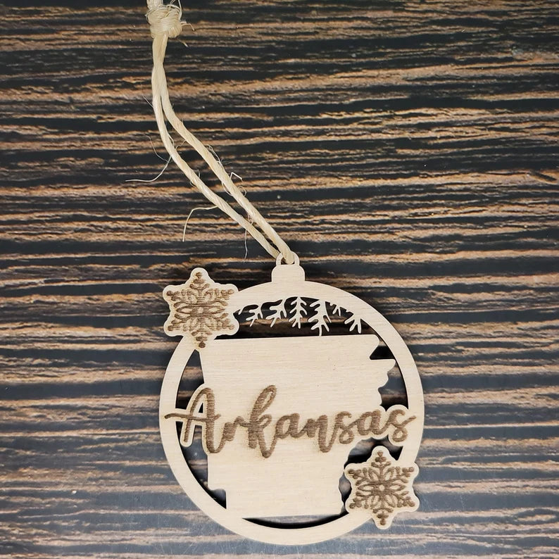 A rustic holiday decor piece, this Arkansas State Christmas Ornament - Laser Engraved Birch Wood by Insanely Paracord is laser engraved from birch wood. It features the outline of Arkansas with trees and the word "Arkansas" in the center, flanked by two carved snowflakes within a round frame. The ornament hangs elegantly from a piece of twine.