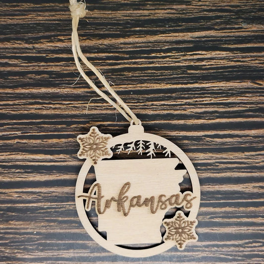 A rustic holiday decor piece, this Arkansas State Christmas Ornament - Laser Engraved Birch Wood by Insanely Paracord is laser engraved from birch wood. It features the outline of Arkansas with trees and the word "Arkansas" in the center, flanked by two carved snowflakes within a round frame. The ornament hangs elegantly from a piece of twine.