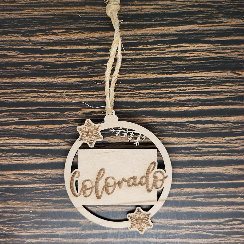 A round wooden Colorado State Christmas Ornament - Laser Engraved Birch Wood, 3" Tall, Natural Twine by Insanely Paracord with "Colorado" laser engraved in decorative cursive. The ornament features a small rectangular shape within a circular border and two carved snowflakes. Hanging from rustic twine against a woodgrain background, it's perfect for rustic home décor.
