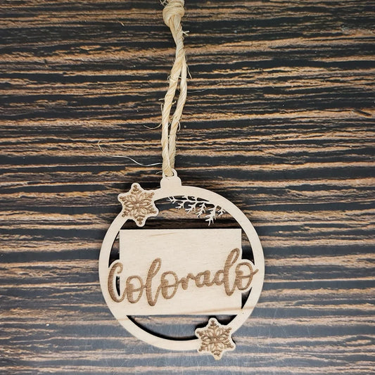 A round wooden Colorado State Christmas Ornament - Laser Engraved Birch Wood, 3" Tall, Natural Twine by Insanely Paracord with "Colorado" laser engraved in decorative cursive. The ornament features a small rectangular shape within a circular border and two carved snowflakes. Hanging from rustic twine against a woodgrain background, it's perfect for rustic home décor.