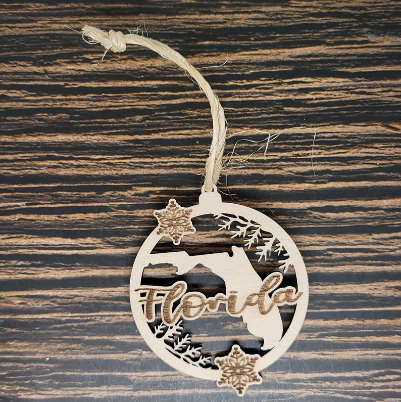 An Insanely Paracord Florida State Ornament - Laser Engraved Birch Wood with Natural Twine features a cutout of the state of Florida with the word "Florida" inscribed beneath it. Snowflakes and pine branches decorate the circular border, crafted from laser-engraved birch wood, and it is hanging by a rustic twine loop on a wooden surface.