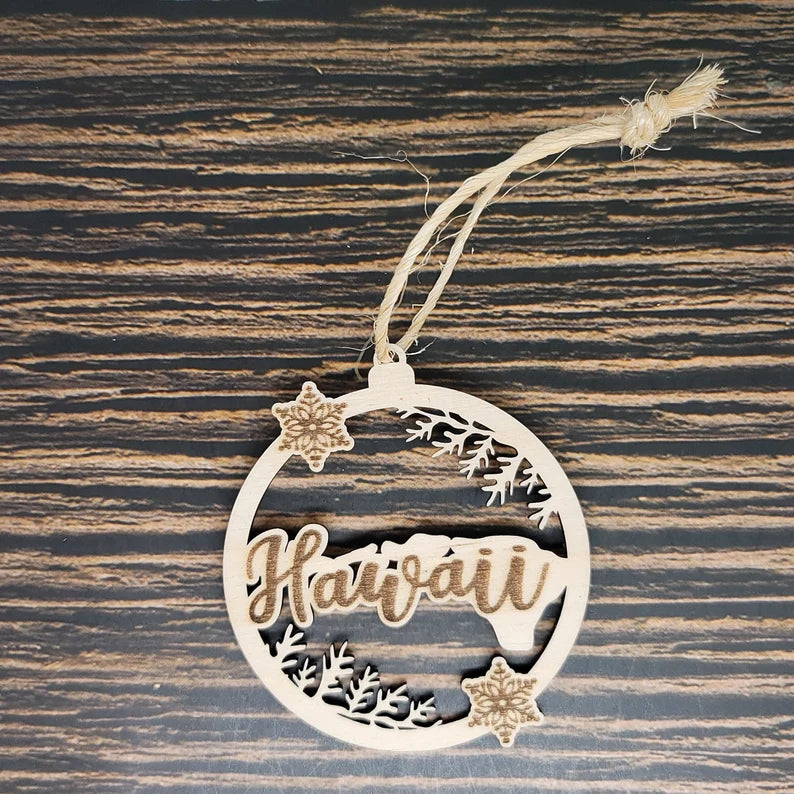 A round birch wood ornament with the word "Hawaii" intricately laser engraved in the center. The design includes small floral shapes and patterns around the circle. A piece of twine is tied at the top for hanging. The background is a dark, striped wood surface. Perfect as an Insanely Paracord Hawaii Christmas Ornament - 3" Tall Laser Engraved Birch Wood.