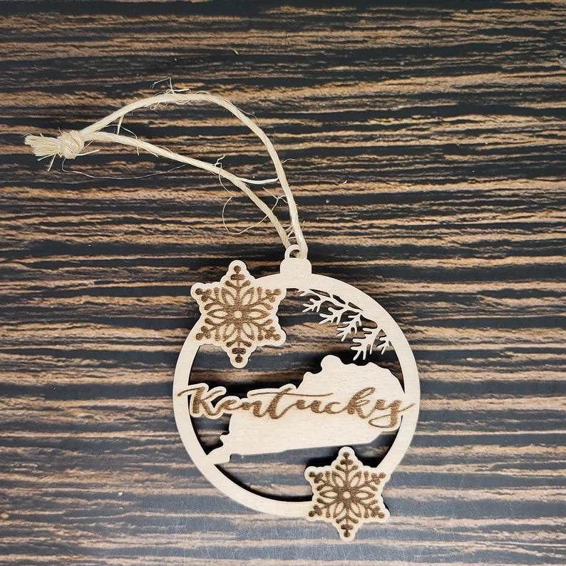 A beautifully handcrafted holiday decoration, this wooden Kentucky State Laser Engraved Christmas Ornament - 3" Tall & 2.75" Wide by Insanely Paracord features "Kentucky" laser engraved across the outline. Adorned with two snowflakes and a sprig of pine, it hangs gracefully by a thin string against a wood-grain background.