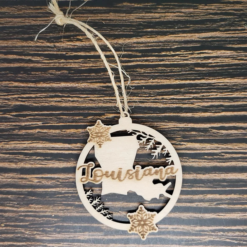An Insanely Paracord Louisiana Laser Engraved Wood Christmas Ornament - 3" Tall, 2.75" Wide with the word "Louisiana" on it. The circular ornament features cutout designs, including a laser-engraved silhouette of Louisiana, snowflakes, and pine branches. It radiates Southern charm as it rests on a dark, striped wooden surface.