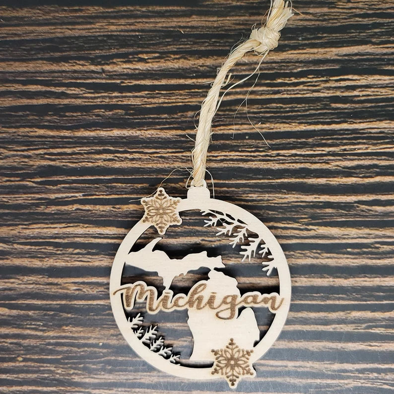 A round wooden ornament displaying the word "Michigan" in cursive, with a silhouette of the state and snowflakes. The Michigan State Christmas Ornament | Laser Engraved Birch Wood by Insanely Paracord hangs from rustic twine against a wooden background, making it perfect holiday decor.