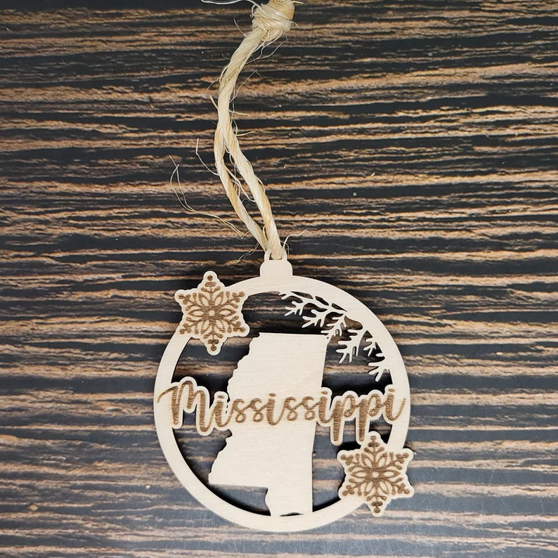 An Insanely Paracord Mississippi Christmas Ornament - Laser Engraved Birch Wood, 3" Tall & 2.75" Wide, with "Mississippi" in cursive. It features the silhouette of the state with two snowflakes and a small pine branch. Suspended by a piece of twine, this ornament is perfect holiday decor.