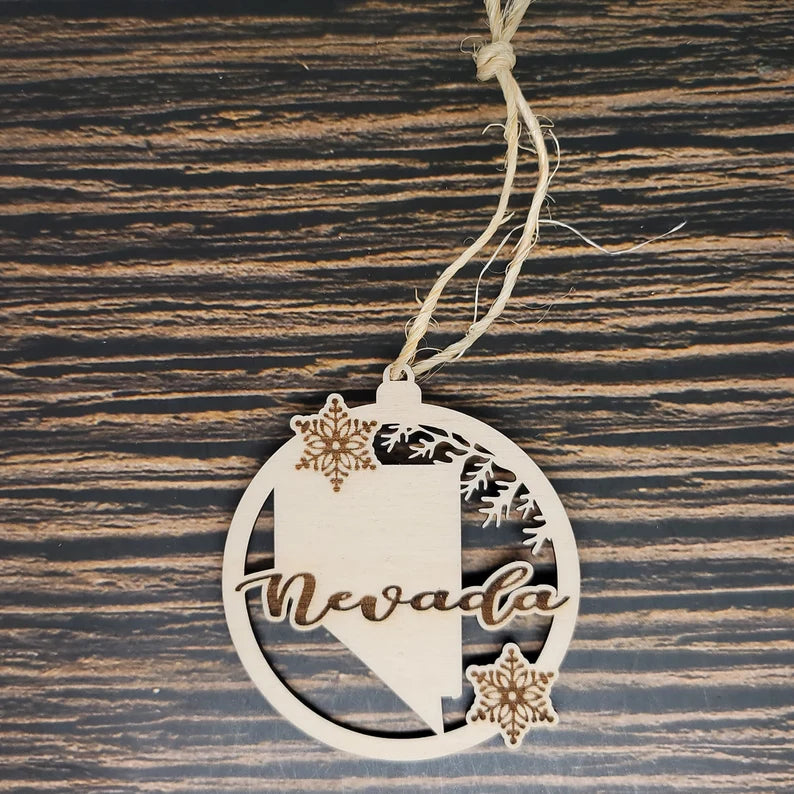 The Nevada State Laser Engraved Wood Christmas Ornament by Insanely Paracord features the word "Nevada" elegantly engraved in cursive. This circular piece features a detailed laser engraving of the Nevada state outline and two delicate snowflakes. Attached to a string for easy hanging, its textured wood background adds rustic charm.