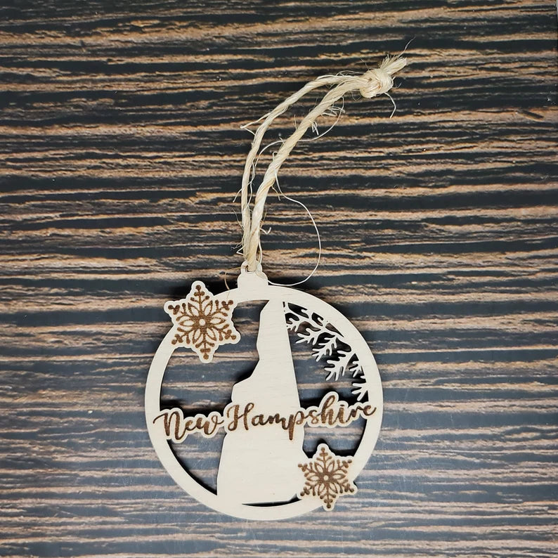 An Insanely Paracord New Hampshire Laser Engraved State Christmas Ornament with intricate carvings, featuring the state's silhouette and two snowflakes, is hanging by a twine loop. This holiday décor piece rests against a dark wood surface with horizontal stripes, adding charm to your 50 states ornaments collection.