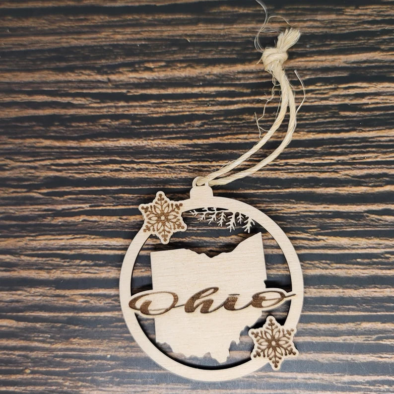 An Ohio State Laser Engraved Wood Christmas Ornament - 3" Tall, 1/8" Birch Wood, Natural Twine by Insanely Paracord features the outline of Ohio with "Ohio" elegantly laser-engraved across it. The circular Ohio State ornament is adorned with two intricate snowflake designs. It hangs from a piece of twine and rests on a striped wooden surface.