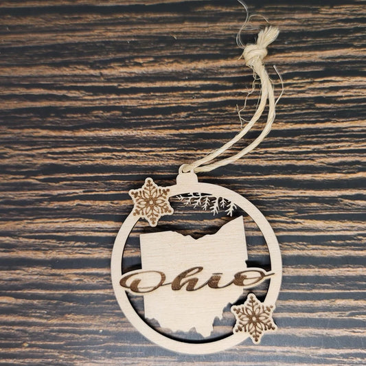 An Ohio State Laser Engraved Wood Christmas Ornament - 3" Tall, 1/8" Birch Wood, Natural Twine by Insanely Paracord features the outline of Ohio with "Ohio" elegantly laser-engraved across it. The circular Ohio State ornament is adorned with two intricate snowflake designs. It hangs from a piece of twine and rests on a striped wooden surface.