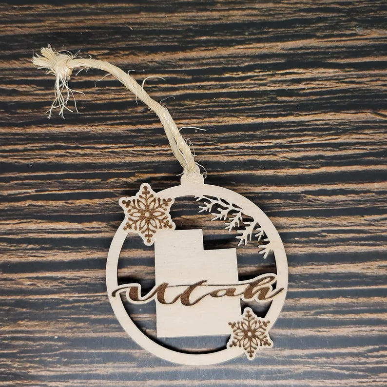 A circular wooden Christmas ornament with the word "Utah" cut out in cursive. It features two intricate snowflake designs and a silhouette of the state of Utah, all expertly laser engraved. The Utah State Laser Engraved Wood Christmas Ornament by Insanely Paracord hangs by a rustic twine string against a dark, textured wood background.