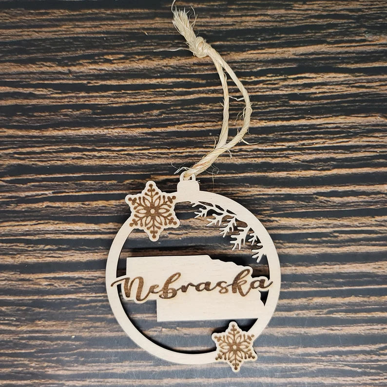 A round, laser-engraved Birch wood ornament with the word "Nebraska" cut out, featuring the shape of the state and two snowflake designs. This Nebraska State Christmas Ornament - Laser Engraved Birch Wood from Insanely Paracord hangs on a string against a dark wooden background, perfect holiday decor for any Nebraskan home.