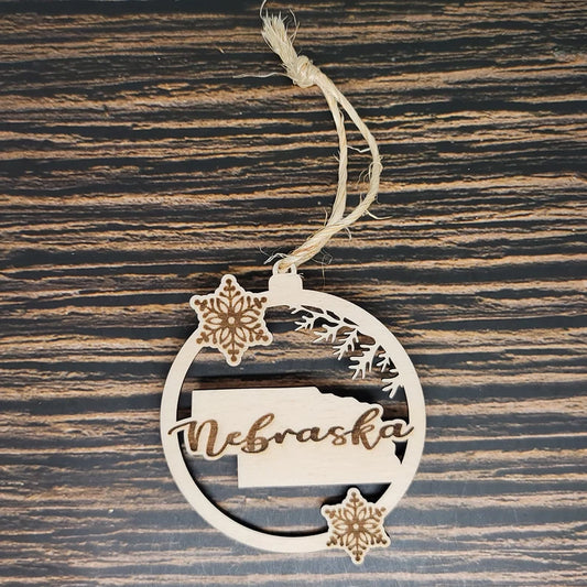 A round, laser-engraved Birch wood ornament with the word "Nebraska" cut out, featuring the shape of the state and two snowflake designs. This Nebraska State Christmas Ornament - Laser Engraved Birch Wood from Insanely Paracord hangs on a string against a dark wooden background, perfect holiday decor for any Nebraskan home.