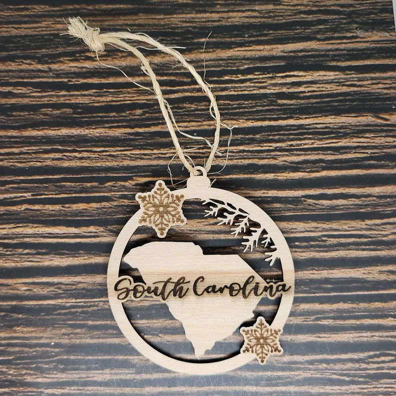 An Insanely Paracord South Carolina Christmas Ornament - Laser Engraved Birch Wood, 3" Tall featuring the state's outline and the words "South Carolina" engraved. The birch wood ornament is decorated with small carved snowflakes and pine branches, all laser engraved. It hangs from a rustic twine loop against a patterned wooden background.