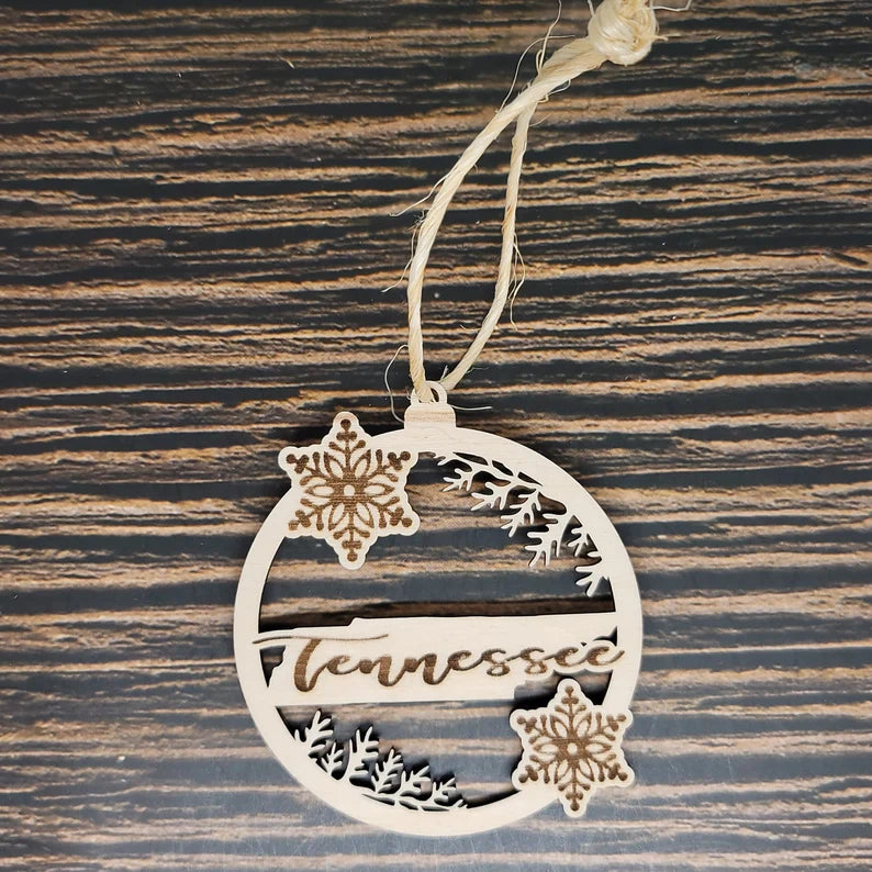 An Insanely Paracord Tennessee Laser Engraved State Christmas Ornament made from birch wood features intricate cut-out designs, including two snowflakes and the word "Tennessee" in cursive, hanging on a knotted string against a dark woodgrain background.