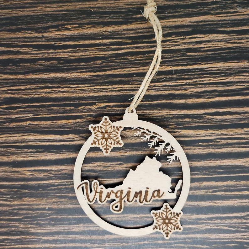 A round wooden ornament features the word "Virginia" with an outline of the state and two carved snowflakes. The Insanely Paracord Virginia Laser Engraved Christmas Ornament - 3" Tall Birch Wood is displayed against a wood grain background, hanging from a twine loop, making it perfect holiday decor.