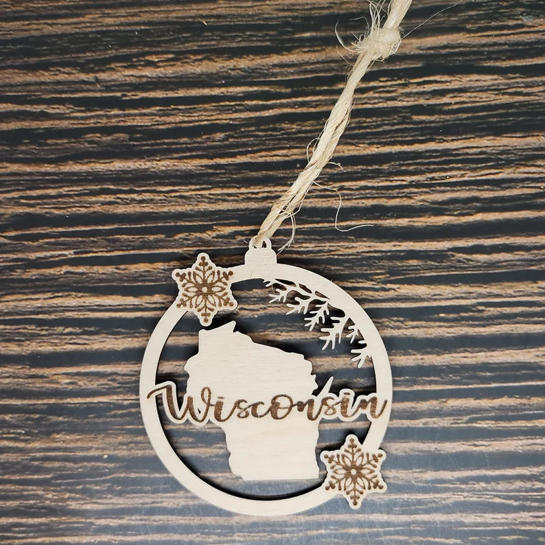 A Wisconsin State Christmas Ornament - Laser Engraved Birch Wood - 3" Tall, 2.75" Wide by Insanely Paracord featuring an outline of Wisconsin in the center with snowflakes above and below. The word "Wisconsin" is cut out in a script font across the state outline. This holiday decor piece hangs from a rustic twine loop, resting on a dark wood background.