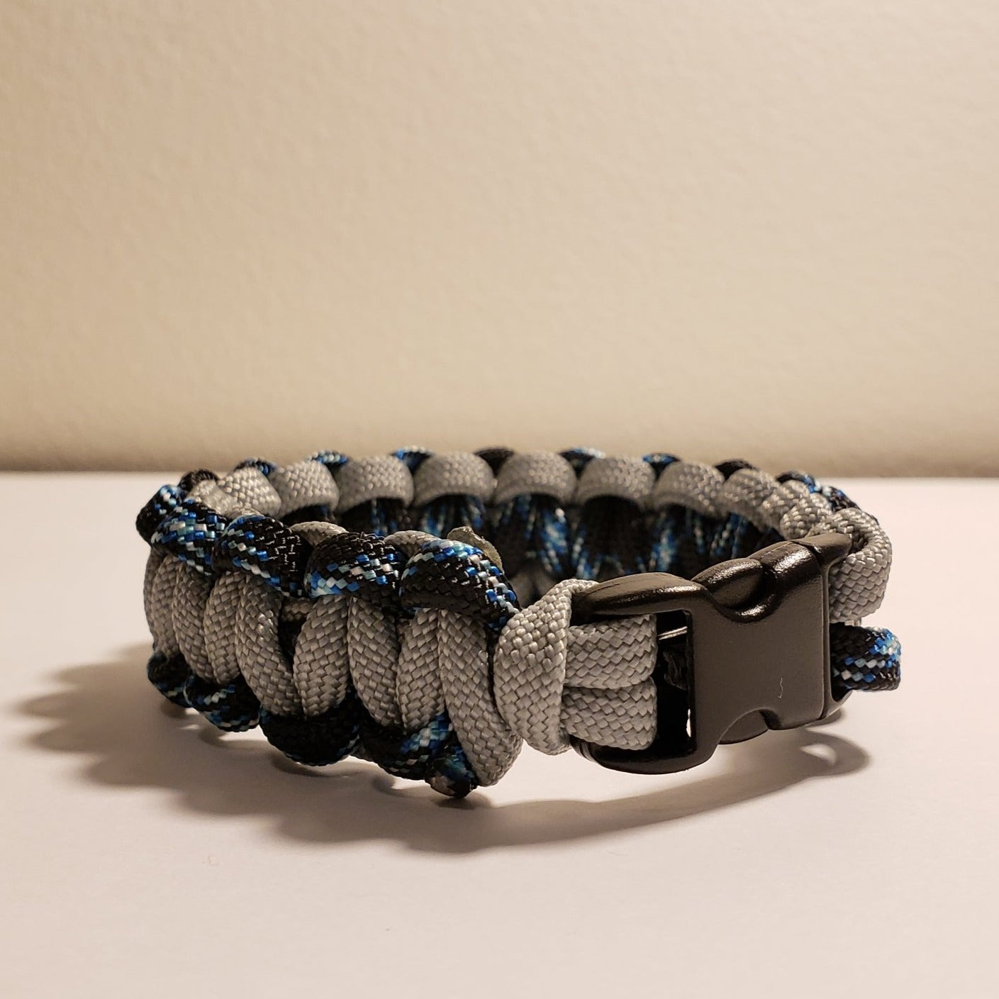A gray, black, and blue custom paracord bracelet with a black plastic buckle is displayed against a plain light-colored background. Handmade in the USA, this Insanely Paracord Custom Paracord Bracelet - 2 Color Choices features a cobra knot pattern and serves as a versatile survival tool.