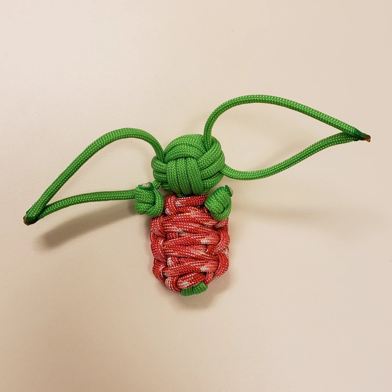 A small decorative knot made from red and green paracord, resembling a strawberry with green leaves. The green cord forms a double loop at the top, resembling the knot’s stem and leaves, while the red cord is intricately woven to represent the body of the strawberry—a charming example of Green Alien Baby - Handmade Paracord Alien Doll - Mint & Strawberry - 550 Paracord - Made in USA by Insanely Paracord creations.