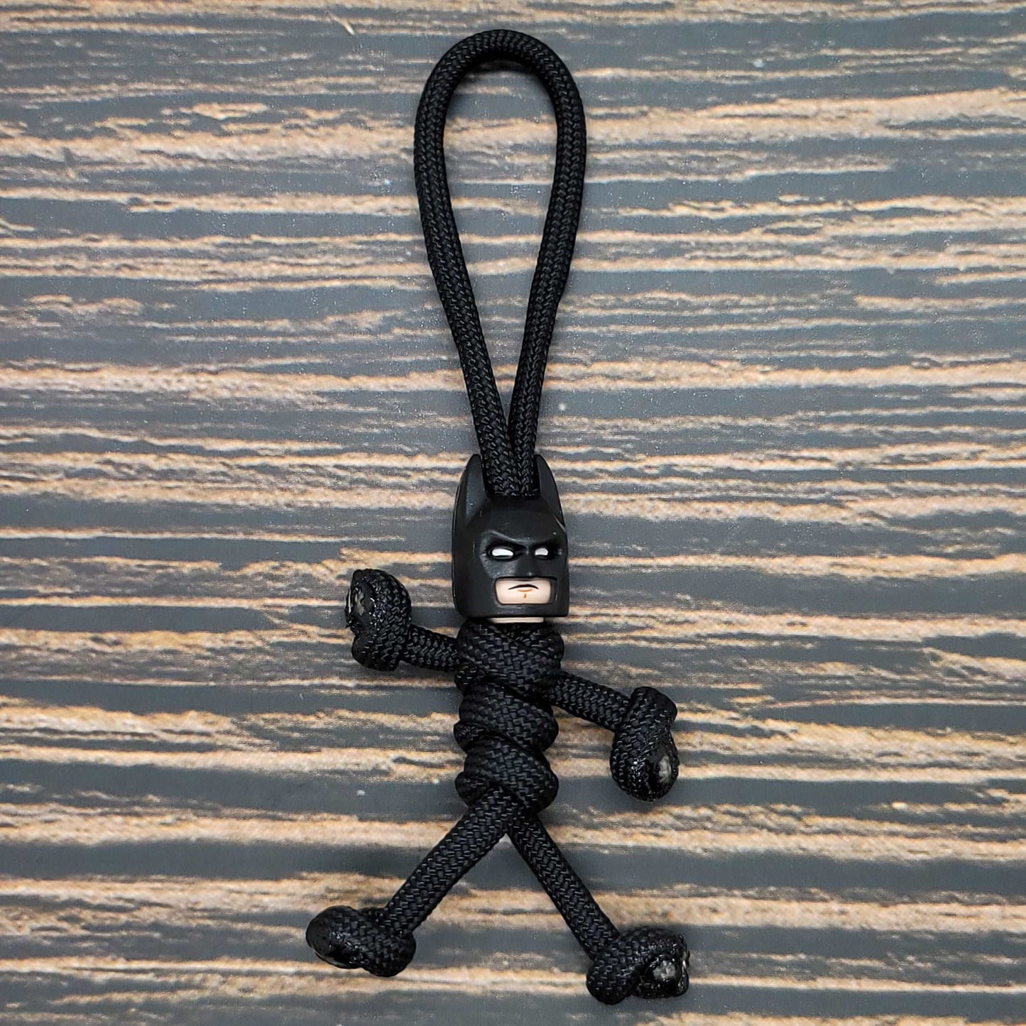 A small figurine shaped like Batman made from black 550 paracord with a plastic Batman head. This handcrafted in the USA Insanely Paracord Bat Super Hero Paracord Pal Keychain Paracord Buddy features knotted arms and legs, creating a human-like form, and is placed on a striped wooden surface.