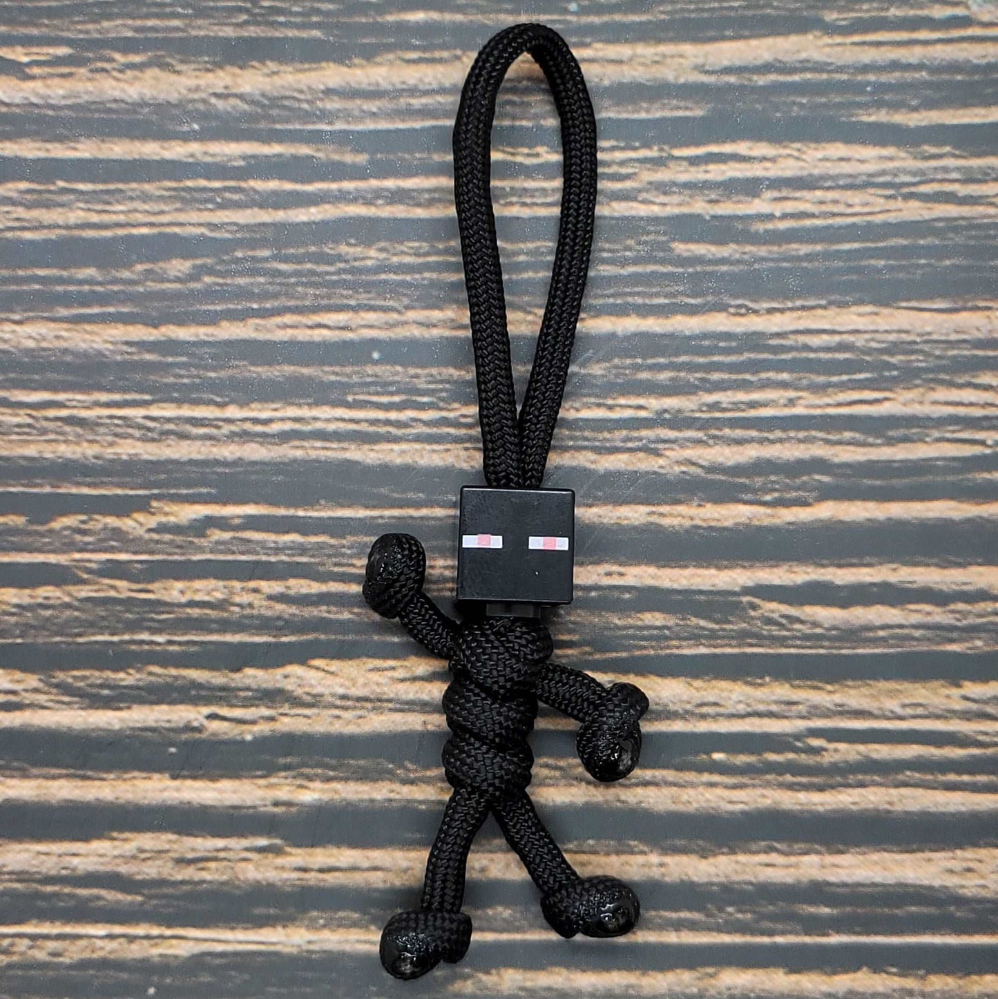 A small black Insanely Paracord Paracord Pal Keychain - Handmade Minifigure Head Design shaped like a human figure with a Minifigure Head design. The head has two small pink and white rectangular eyes, and the keychain is placed on a textured wooden surface.