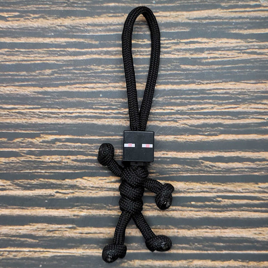 A small black Insanely Paracord Paracord Pal Keychain - Handmade Minifigure Head Design shaped like a human figure with a Minifigure Head design. The head has two small pink and white rectangular eyes, and the keychain is placed on a textured wooden surface.