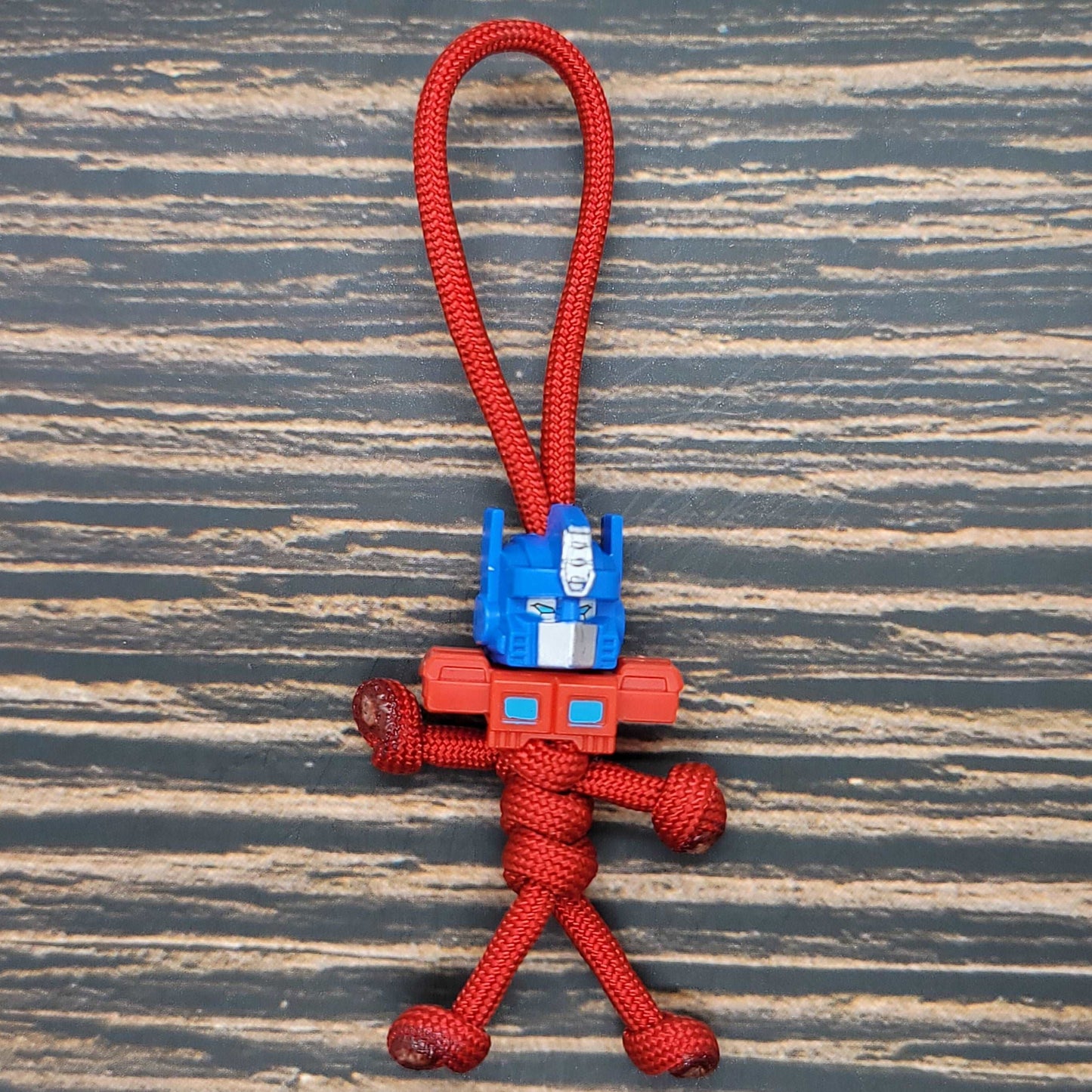A small Car Transformer Semi - 550 Paracord Pal Keychain | Handmade | USA-Made | Red | Mini Figure from Insanely Paracord is designed to look like a robot with a detailed blue and red head. The figurine has a loop at the top for hanging, making it perfect for attaching to your bag or keys. It rests on a striped surface, reminiscent of an Insanely Paracord Car Transformer Semi theme.